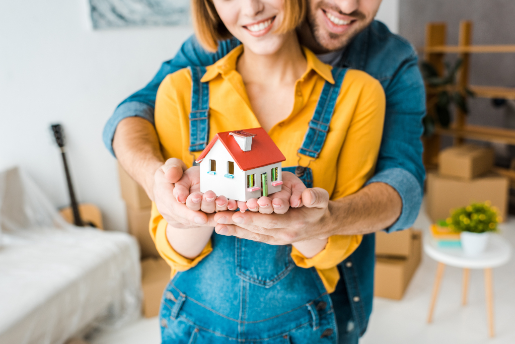 Cropped view of smiling couple holding toy house and looking at camera at home